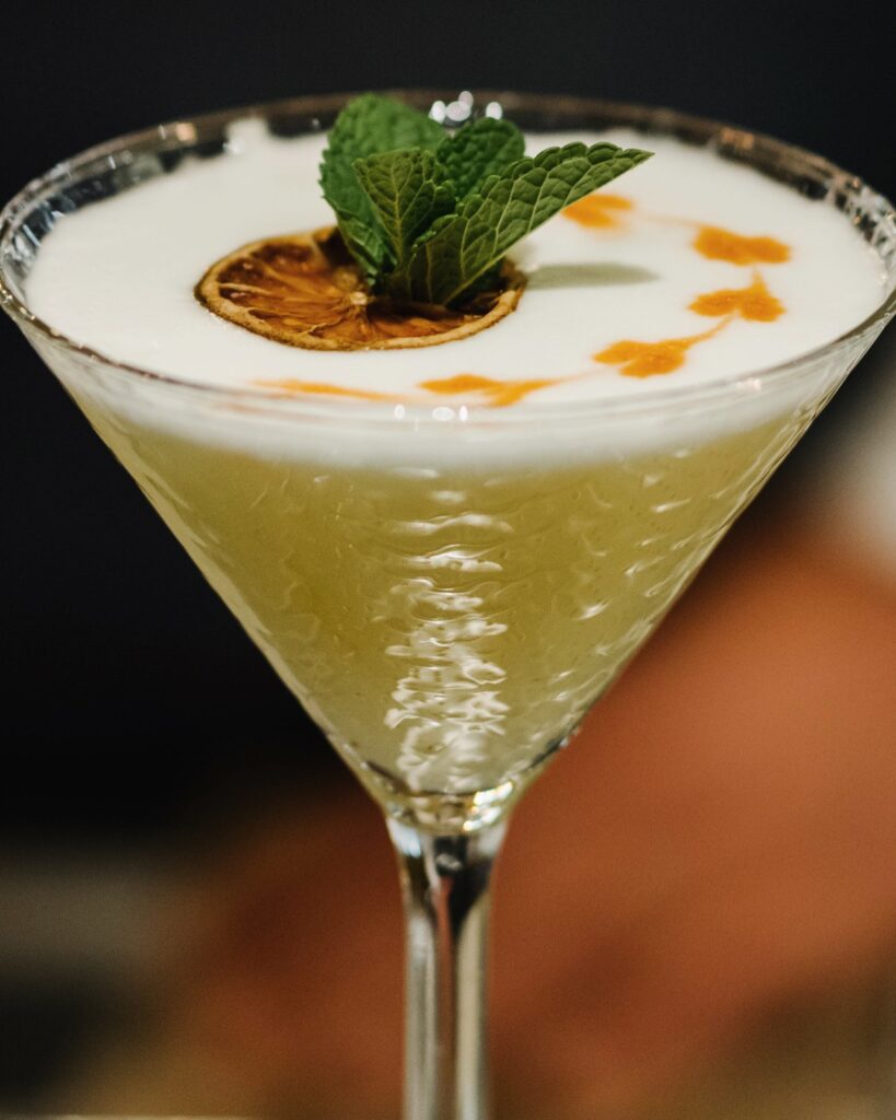 Cocktail premium en verre martini avec mousse blanche, gouttes orange et garniture d’agrumes et menthe