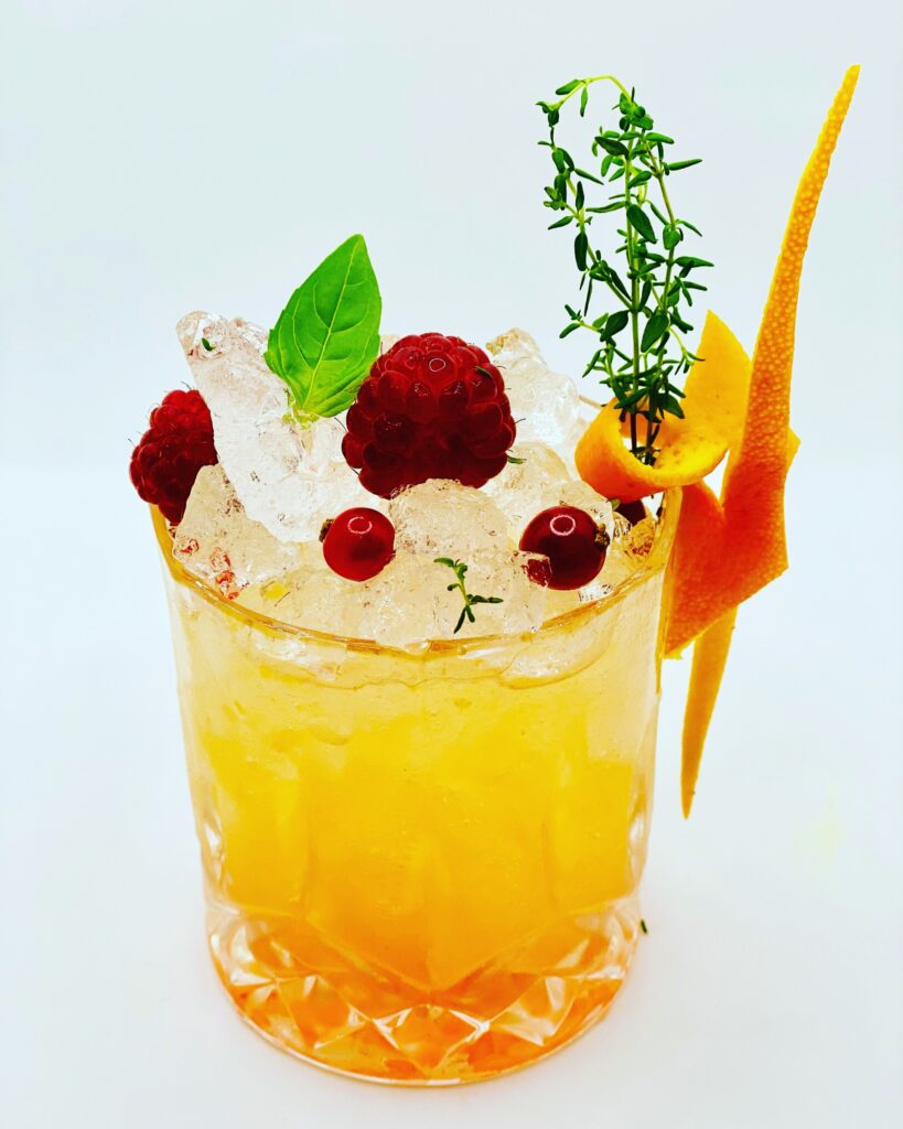Cocktail premium orange-jaune garni de framboises, cranberries, basilic et zeste d’orange